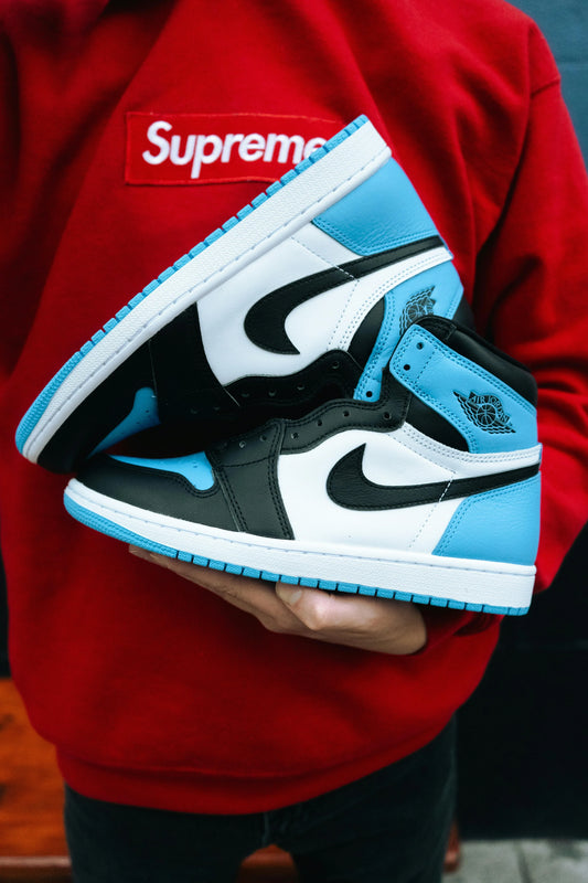 Person wearing a red Supreme sweatshirt holding a pair of blue and black Nike Air Jordan sneakers, showcasing iconic pieces from luxury streetwear brands.