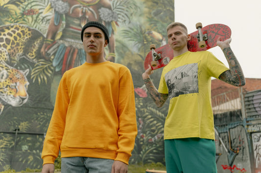 Two men in streetwear clothing posing outdoors; one wearing a yellow sweatshirt and the other holding a skateboard, showcasing streetwear style and fashion.