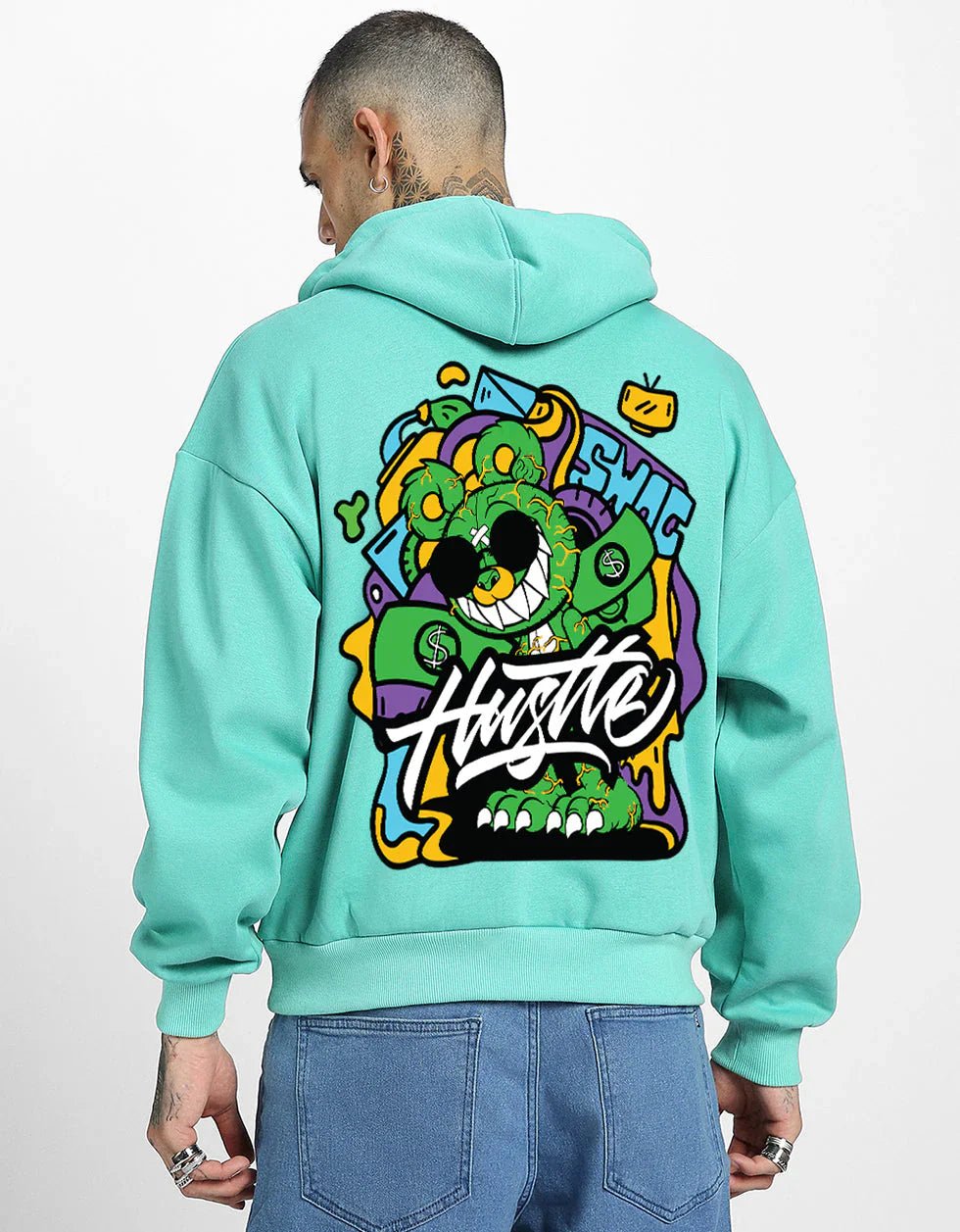 Person wearing a turquoise graphic hoodie featuring a colorful cartoon bear design with sunglasses, graffiti-style text that says "Hustle," and vibrant street art elements.