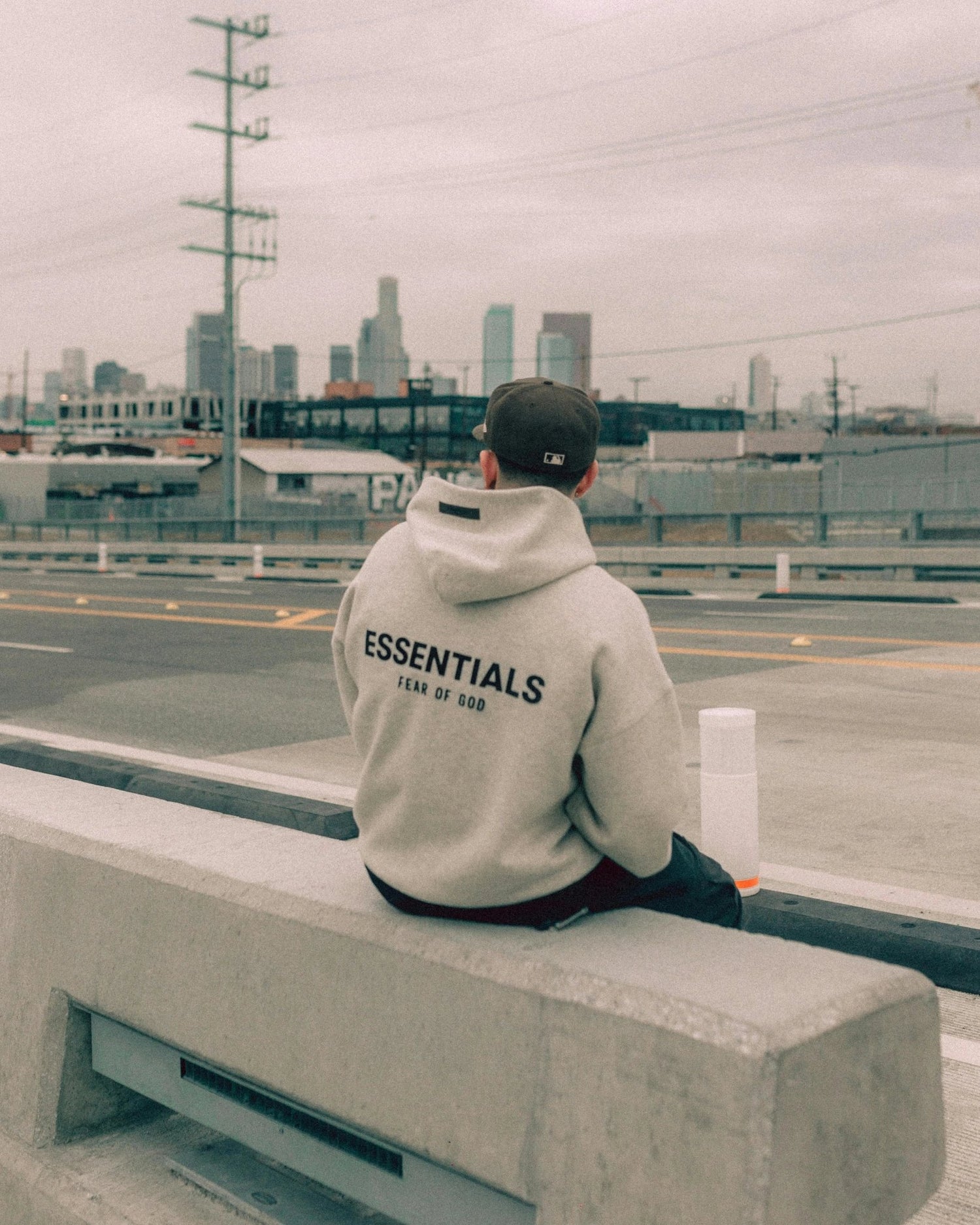 Person wearing a Fear of God Essentials hoodie sitting on a concrete bench overlooking an urban cityscape, showcasing the back logo of the hoodie.