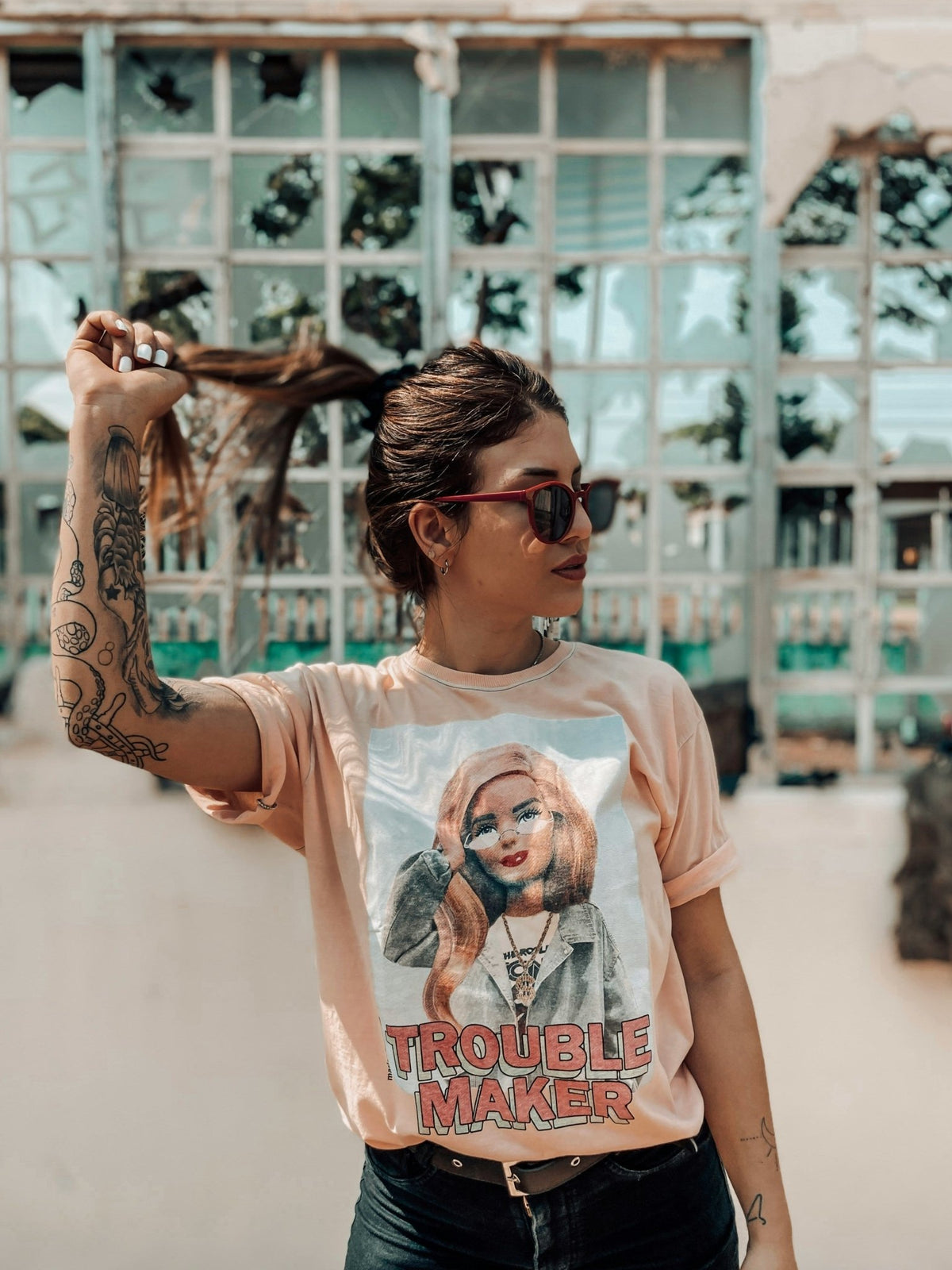 Woman wearing a pink oversized streetwear t-shirt with bold graphic and "Troublemaker" text, styled with sunglasses and tattoos in an urban setting.
