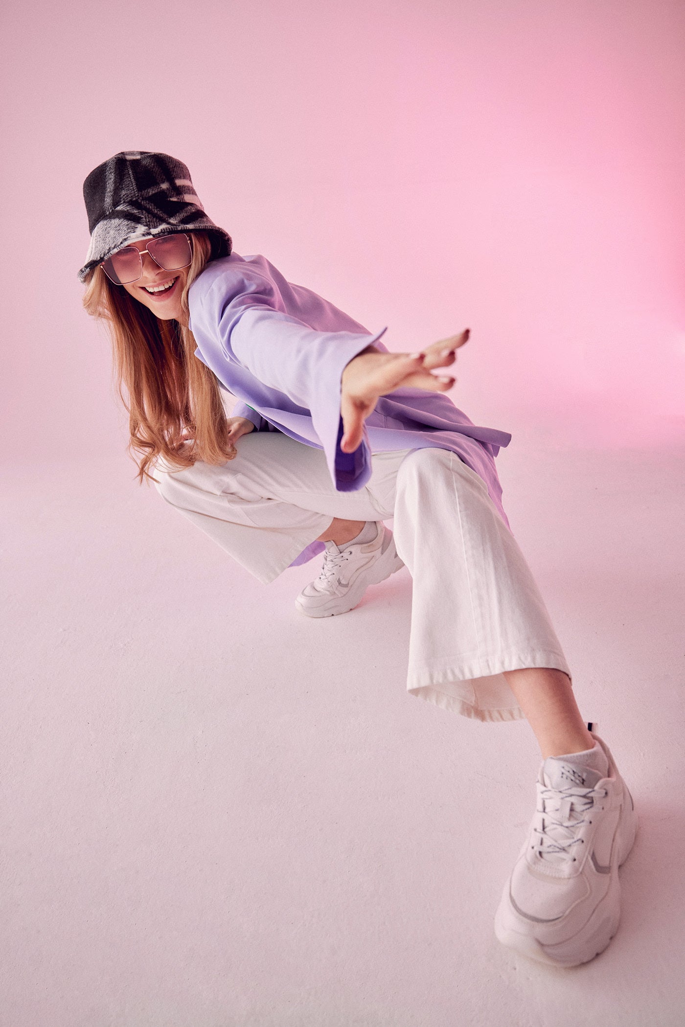 Fashionable young woman wearing a lavender blazer, white track pants, chunky white sneakers, and a stylish plaid bucket hat, crouching playfully in a pink-toned studio setting.