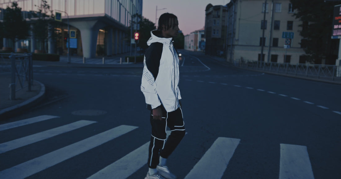A man wearing a white and black reflective men jacket and matching pants stands on a crosswalk in a quiet urban street at dusk, with buildings and a traffic light in the background.